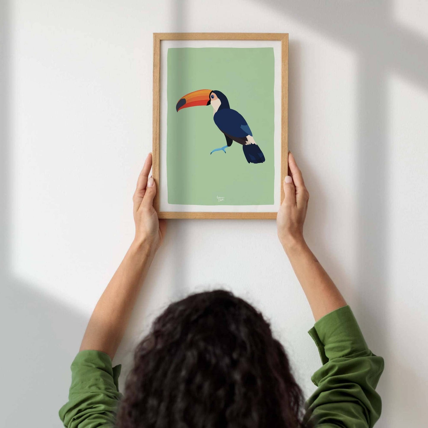 A woman's hands hanging an A4-sized framed toucan art print on a wall. This provides a clear real-life sense of scale, helping customers visualize the perfect size of the A4 print for their gallery wall or bedroom decor.
