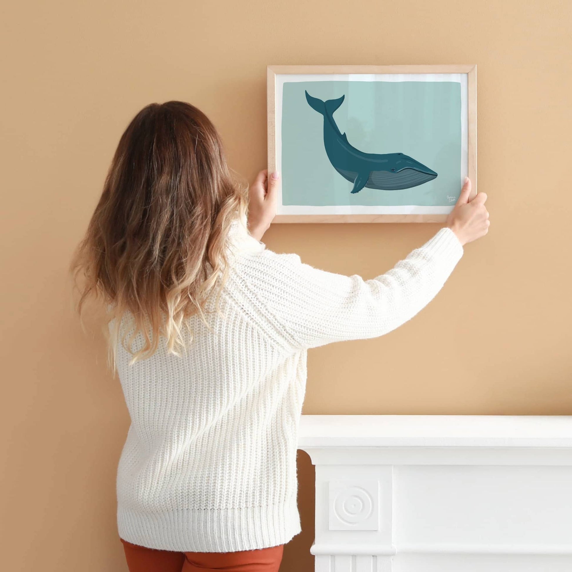 A woman (seen from behind) holding and positioning the framed A4-sized blue whale print on a warm, earthy orange-terra wall, centered over a modern white fireplace mantle. This photo shows how the artwork creates a stunning, contrasting focal point in a curated home interior.