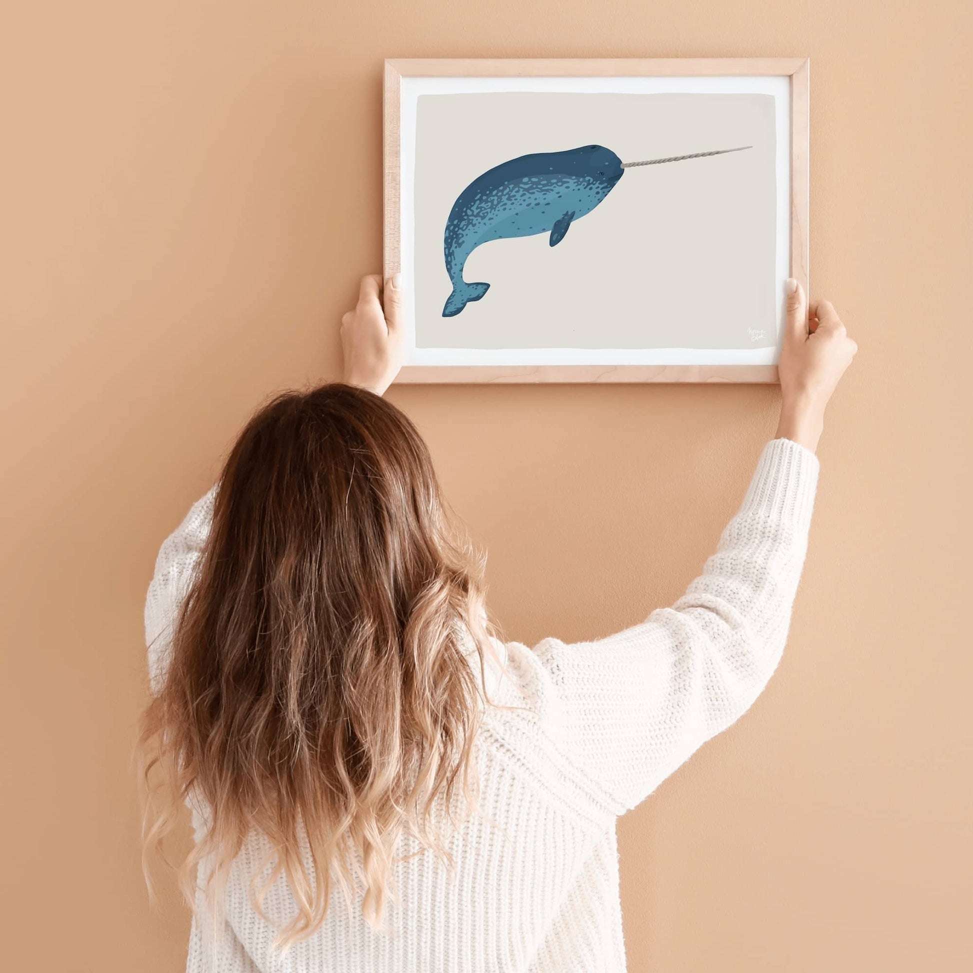 A woman carefully hanging a framed narwhal print on a wall, giving a clear idea of the artwork's scale in a real home setting.