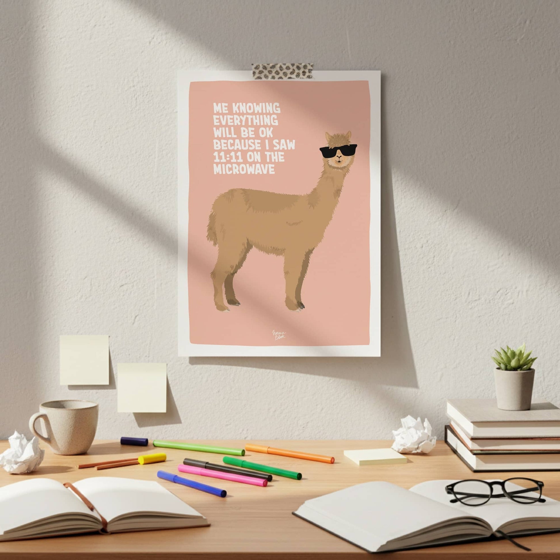 The light coral alpaca poster attached with colorful washi tape to a clean white wall. It hangs directly above a messy office desk with creative clutter, showing a casual, framework-free, and effortless style to decorate an authentic work-from-home setup.