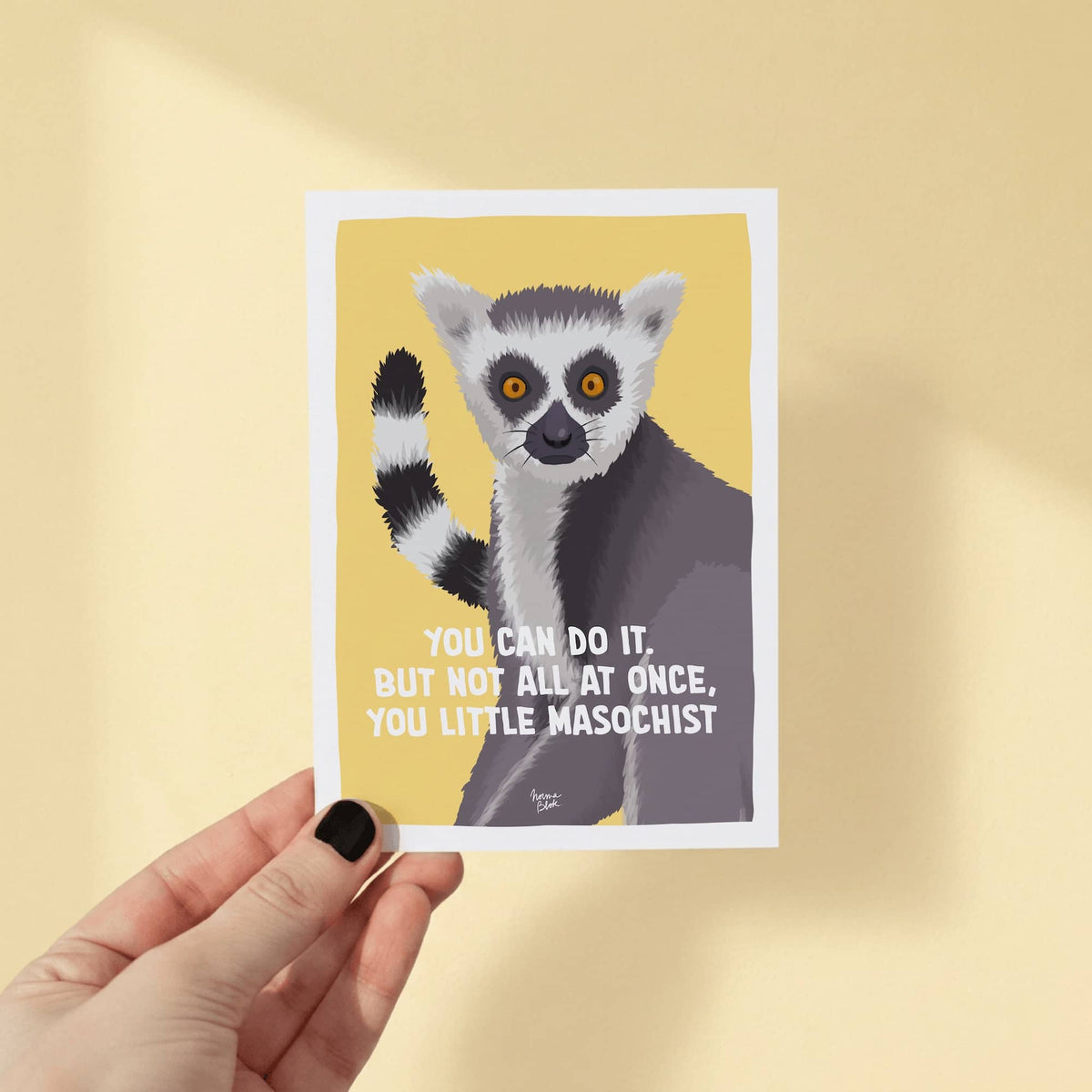 A woman's hand holding an A6 mini print postcard to demonstrate its true physical scale and size against a plain chocolate background. The illustrated card features a wide-eyed ring-tailed lemur staring forward on a bright yellow background with the sarcastic quote "You can do it. But not all at once, you little masochist." in bold typography, showing the clean edges and soft paper texture of the product by Normablok.