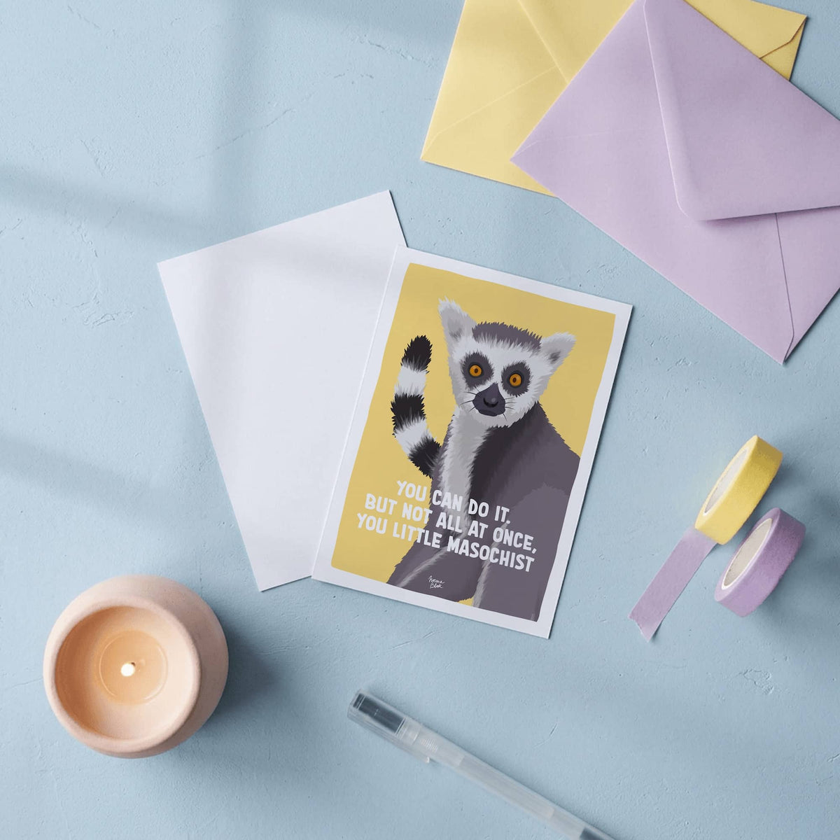 An overhead flat lay display on a clean desk showing two A6 postcards side by side. One postcard shows the front illustrated side with the funny ring-tailed lemur on a yellow background and the quote "You can do it. But not all at once, you little masochist.", while the second postcard is turned face down to clearly show its clean, blank white back designed for writing personal messages or framing by Normablok.